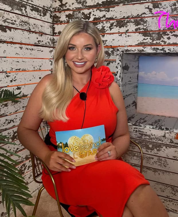 Woman in a red dress holding a 'Love Island' book against a rustic wooden backdrop